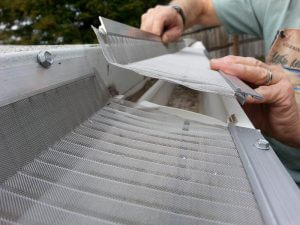 A person installs a mesh gutter guard on a roof edge, securing it to the gutter with metal screws. The guard helps prevent debris from clogging the gutter.