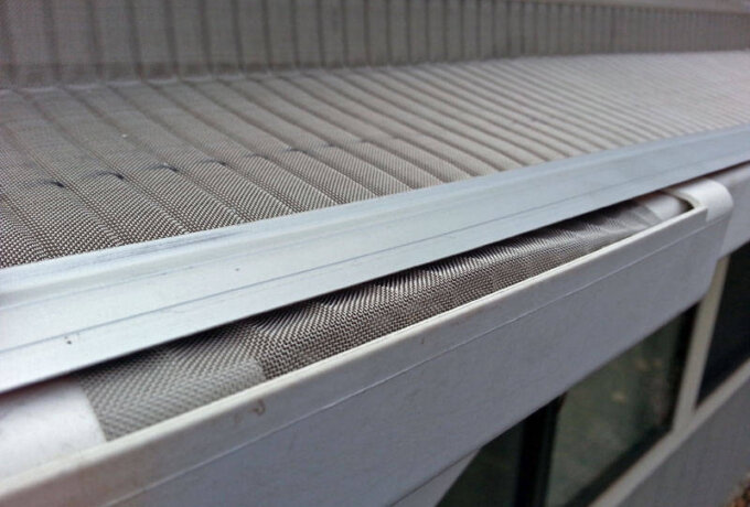 Close-up of a house gutter with a fine mesh cover installed, designed to prevent leaves and debris from entering while allowing water to flow through. The side of the house and a window are partially visible below.