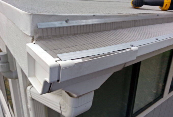 Close-up of a house roof corner with a white gutter guard partially lifted, exposing the gutter below. A screwdriver lies on the roof, and downspouts are attached to the side of the house.
