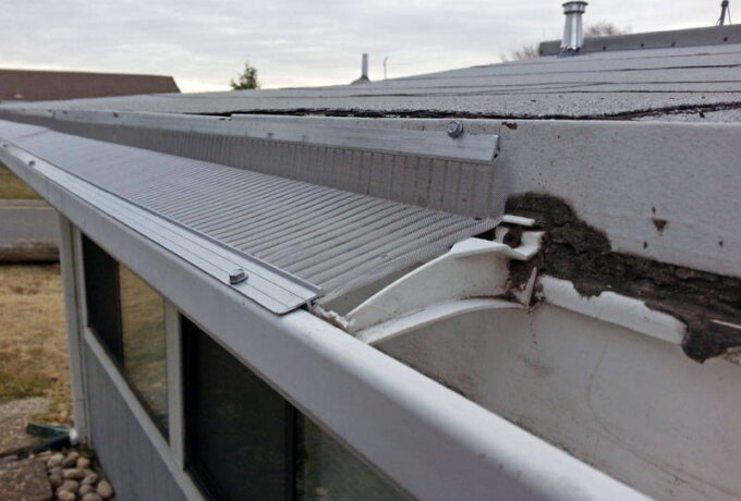 A close-up view of a house’s roof shows damaged gutters and a poorly installed gutter guard, with some debris and cracked concrete visible along the roof edge.