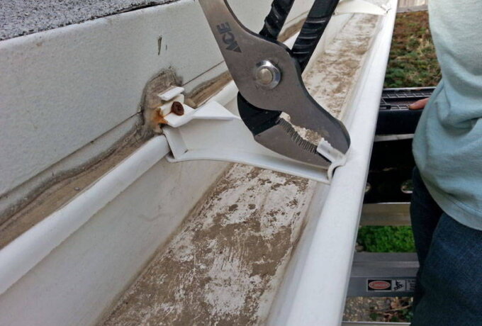 A person using pliers to repair a white plastic gutter bracket attached to a house; the gutter appears dirty and rusty, with the person standing on a ladder.