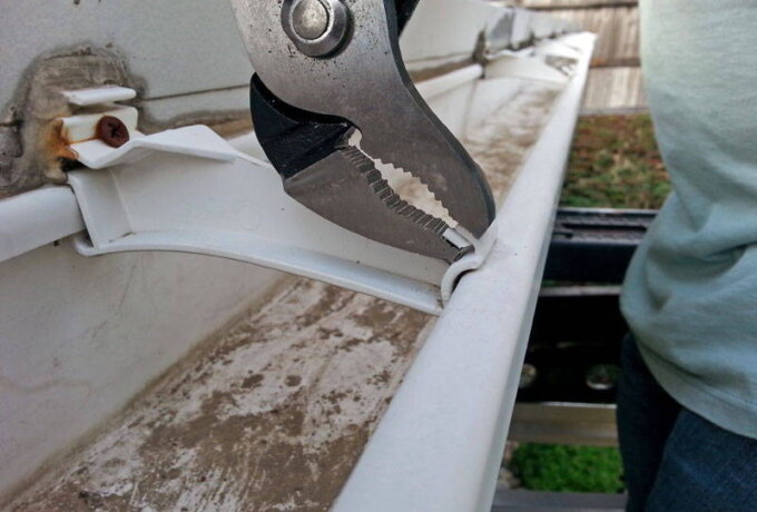 A person uses pliers to repair or pinch together a section of a white, dirty rain gutter attached to a house, with some rust and screws visible nearby.