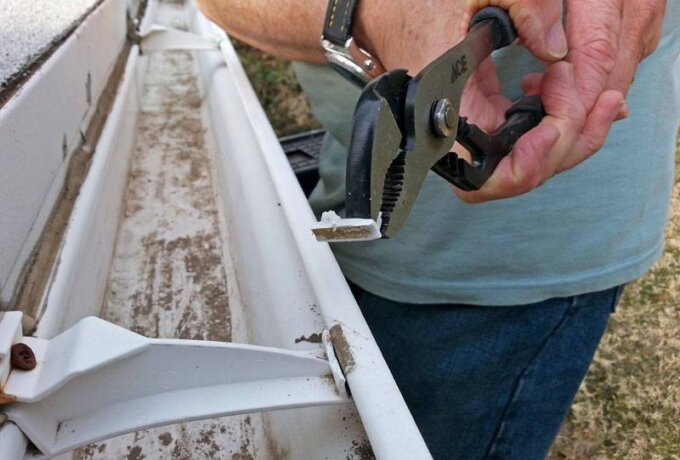 A person uses a pair of pliers to remove debris from a dirty white rain gutter attached to a building.
