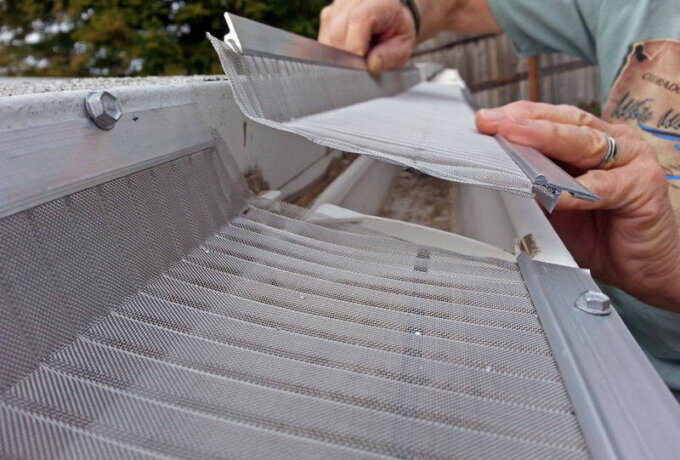 A person installs a metal mesh guard onto a rain gutter, using their hands to secure the screen over the gutter edge, with trees and a fence visible in the background.