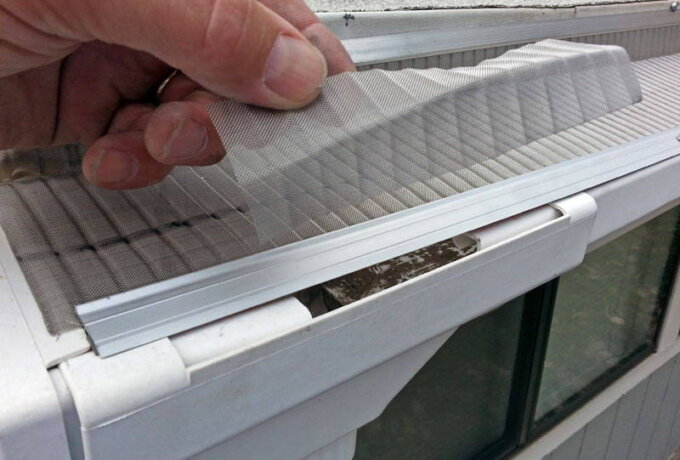 A hand lifts a mesh gutter guard to reveal leaves and debris inside a white rain gutter attached to the edge of a roof above windows.