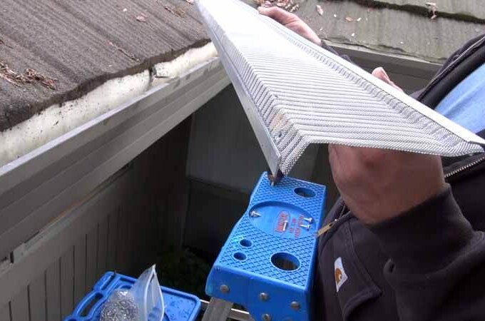 Person standing on a blue ladder near a roof, holding an Easy On Gutter Guard with stainless steel micro-mesh above an open gutter. Roofing shingles and some leaves are visible in the background. Costco quality ensures lasting protection.