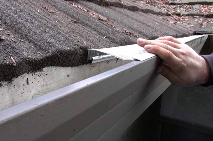A person’s hand is cleaning debris from a rain gutter with leaves and dirt on a sloped roof protected by Easy On Gutter Guard’s stainless steel micro-mesh.