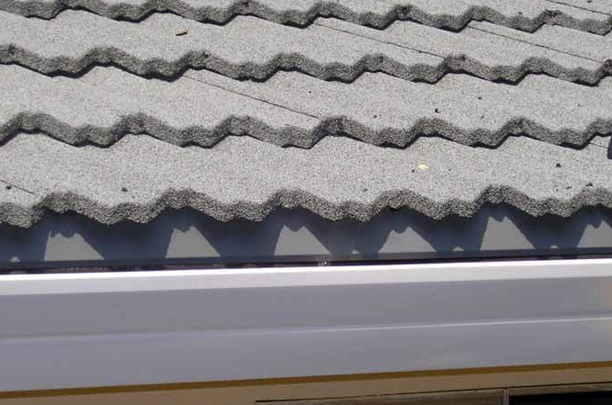 Close-up of a roof with gray, wavy concrete tiles and a white Gutterglove gutter guard running along the edge. Some debris and a small black object are visible on the roof, highlighting its easy installation.
