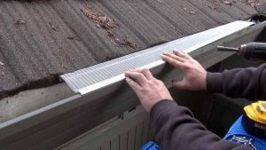 A person installs a metal gutter guard along the edge of a roof covered with brown shingles, using a power drill and measuring tools.
