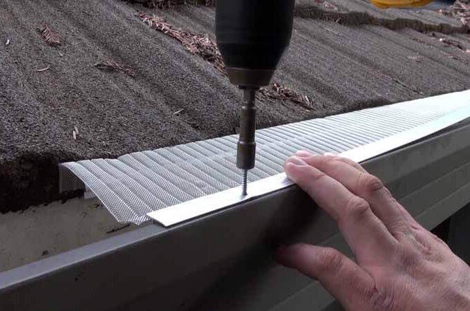 A person uses a drill to attach an Easy On Gutter Guard onto the edge of a roof gutter to prevent debris from entering.