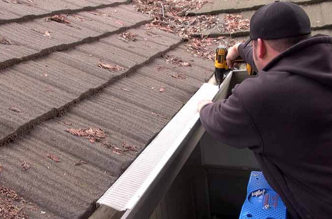 A person in a dark hoodie and beanie uses a power drill to install an Easy On Gutter Guard along the edge of a shingled roof covered with fallen leaves and debris.
