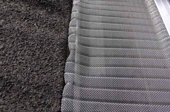 Close-up of a Gutterglove metal mesh gutter guard installed along the edge of a roof with rough, dark asphalt shingles. The mesh offers easy installation and is designed to keep debris out of the gutter. Costco option available.