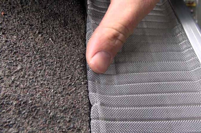 A hand presses down on a stainless steel micro-mesh Gutterglove gutter guard being installed along the edge of a roof with asphalt shingles for easy installation.