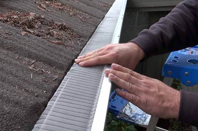 A person installs a Gutterglove mesh gutter guard on the edge of a roof, pressing it into place with both hands. Pine needles and debris are visible on the roof, and a blue step ladder is nearby for easy installation.