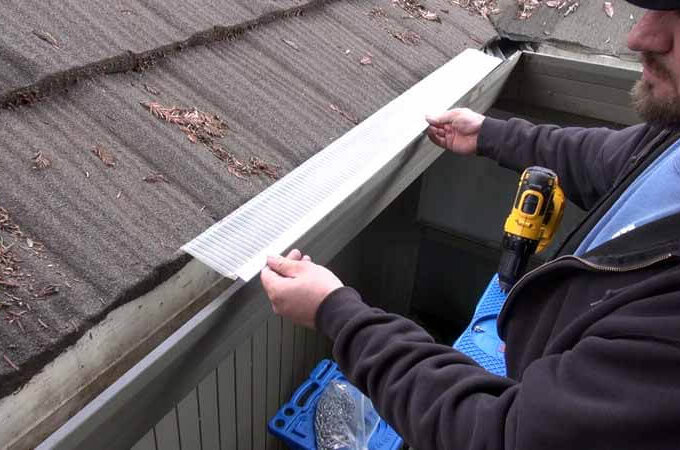 A person standing on a ladder installs a Gutterglove stainless steel micro-mesh gutter guard along the roof edge, with a drill nearby and leaves scattered on the roof shingles.