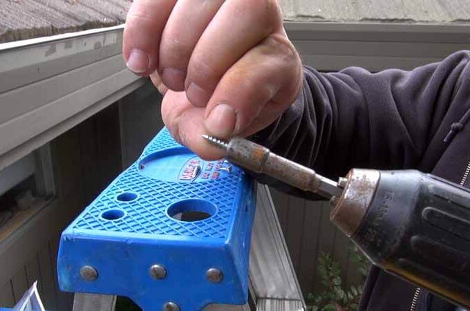 A person on a blue step ladder uses a power drill to drive a screw into place for easy installation, with part of a building visible in the background. Gutterglove products available at Costco may be featured.