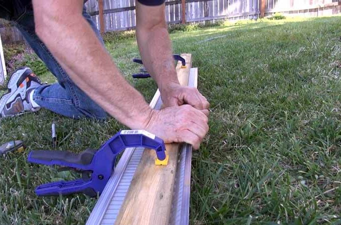 A person kneels on grass, using clamps to secure a piece of wood to a metal guide, preparing for an outdoor woodworking project featuring Gutterglove’s stainless steel micro-mesh for easy installation.