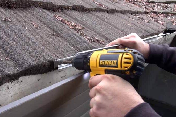 A person uses a yellow DeWalt power drill to fasten or repair a metal gutter with a stainless steel micro-mesh gutter guard, attached to a roof covered in brown shingles and scattered leaves.