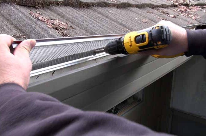 A person uses a yellow power drill to install a Gutterglove stainless steel micro-mesh gutter guard along the edge of a house roof, with some leaves visible on the shingles, highlighting its easy installation.