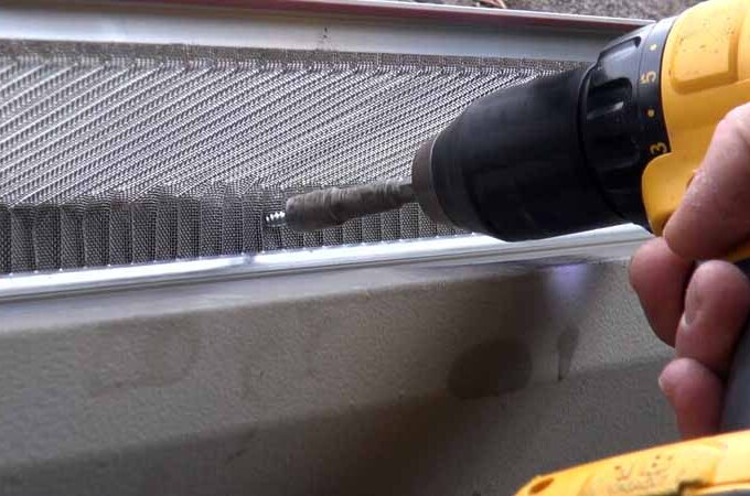 A person uses a yellow power drill to install a Costco stainless steel micro-mesh gutter guard, securing it over a metal mesh screen, possibly on a gutter or vent.