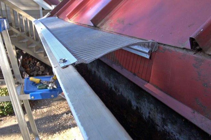 A close-up of a red metal roof and gutter with a partially installed mesh guard, a blue step ladder nearby, and stairs and firewood visible in the background.