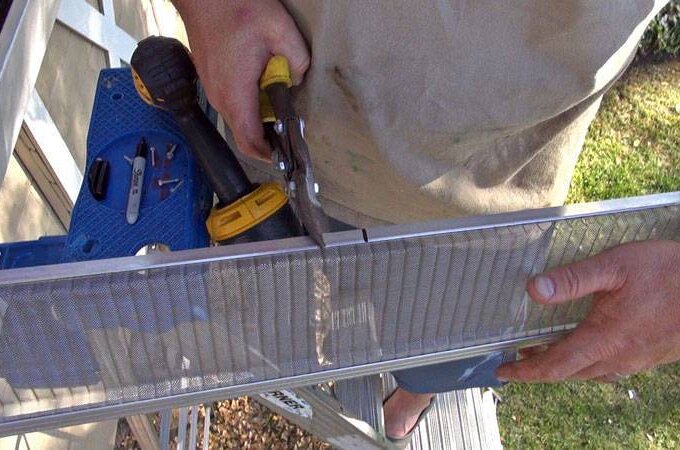 A person uses tin snips to cut a piece of mesh gutter guard while holding a drill, working on a ladder near a house.