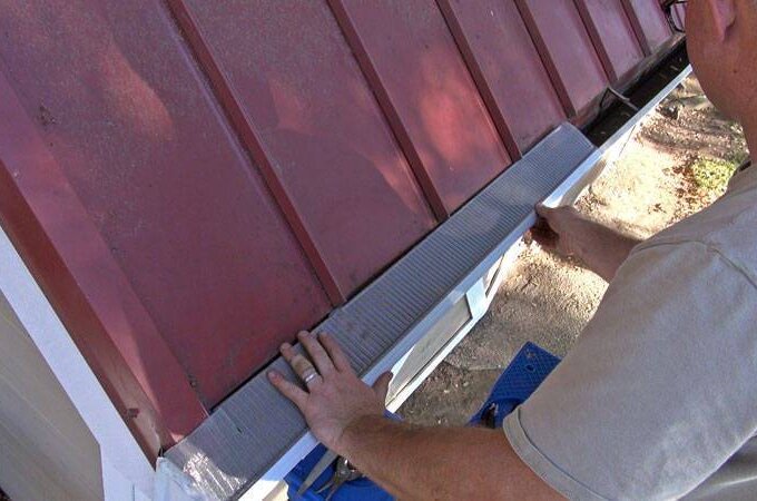 A person installs a piece of metal flashing along the edge of a red metal roof, using both hands to secure it in place near the roofline of a small building.