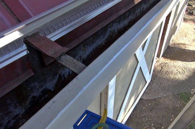View of a house’s roofline showing a clogged rain gutter filled with dark debris and leaves, with a tool and ladder visible, indicating gutter cleaning in progress. The side of the house and yard are also visible.