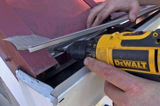 A person uses a yellow DeWalt power drill to attach a metal mesh screen to the edge of a red metal roof above a white structure.