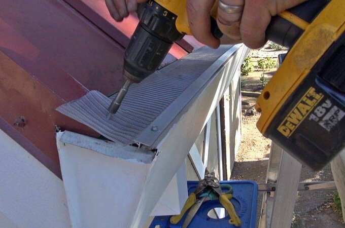 A person uses a DeWalt cordless drill to install a metal mesh gutter guard on the edge of a roof. Tools and a blue platform are visible below the gutter.