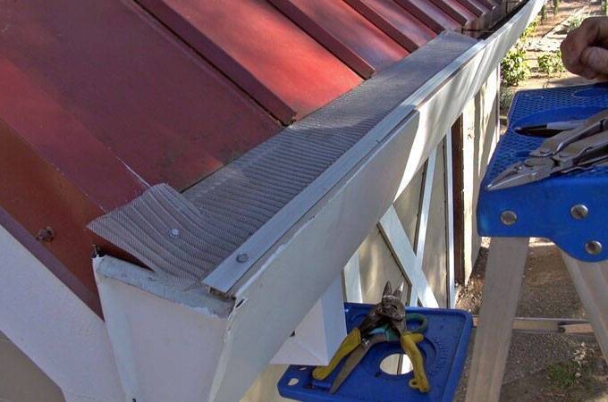 A person stands on a blue ladder installing a metal gutter guard on a red-roofed building. Various tools, including pliers and screwdrivers, are placed on the top of the ladder near the gutter.