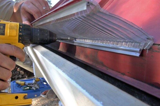 Close-up of a person using a cordless drill to install a mesh gutter guard along the edge of a red metal roof. Tools and materials are visible in the background.