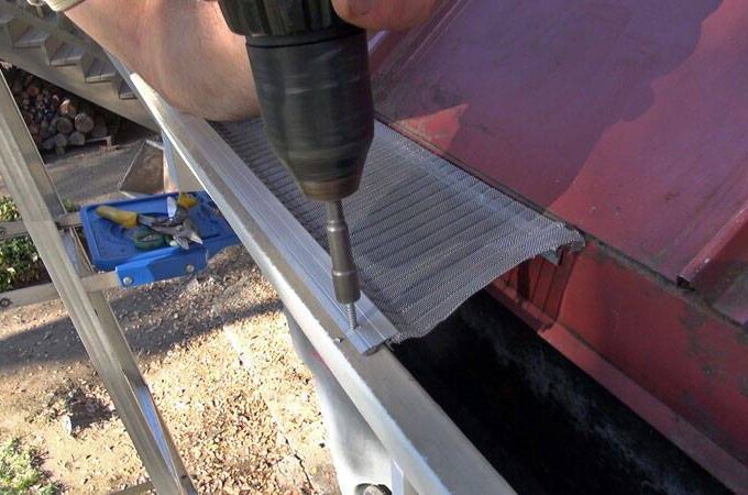 A person uses a power drill to install a mesh gutter guard along the edge of a roof, with a ladder and tools visible nearby.