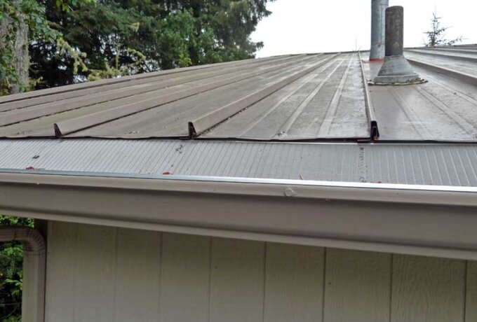 A close-up view of a metal roof with a rain gutter guard installed along the edge; trees and part of a pipe vent are visible in the background.