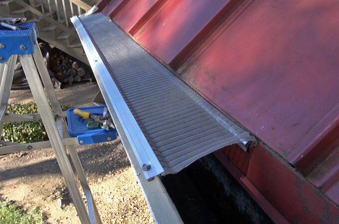 A metal mesh gutter guard is being installed on the edge of a red metal roof. A blue stepladder with tools rests nearby, and the scene is outdoors with grass and wood in the background.