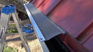A close-up of a red metal roof with a silver mesh gutter guard being installed. A blue step ladder with tools sits nearby, and trees and logs are visible in the background.