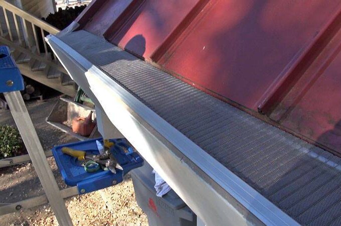 A close-up view of a house roof with a metal gutter guard installed along the edge. A step ladder with tools is set up nearby, and some leaves and debris are visible on the ground below.
