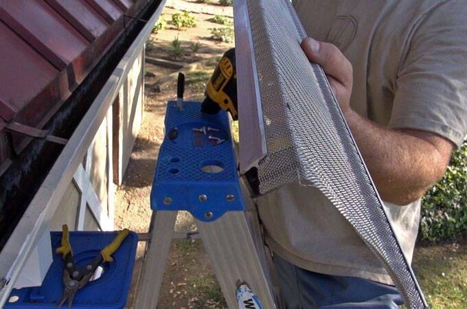 A person standing on a ladder installs a mesh gutter guard on a roof edge, holding a power drill and tools nearby, with a garden visible in the background.