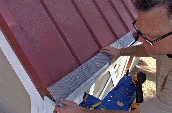 A person wearing glasses installs or adjusts metal flashing along the edge of a red metal roof on a small white shed, using a ladder for support.