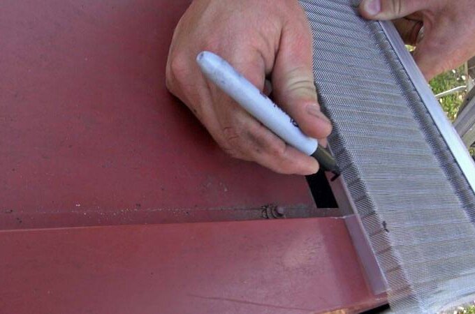 A person uses a black marker to trace the edge of a metal mesh screen onto a red surface, possibly preparing to cut or install the screen.