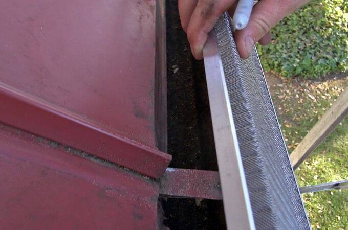 A person is installing a metal gutter guard on the edge of a red metal roof while standing on a ladder, with a yard visible below.