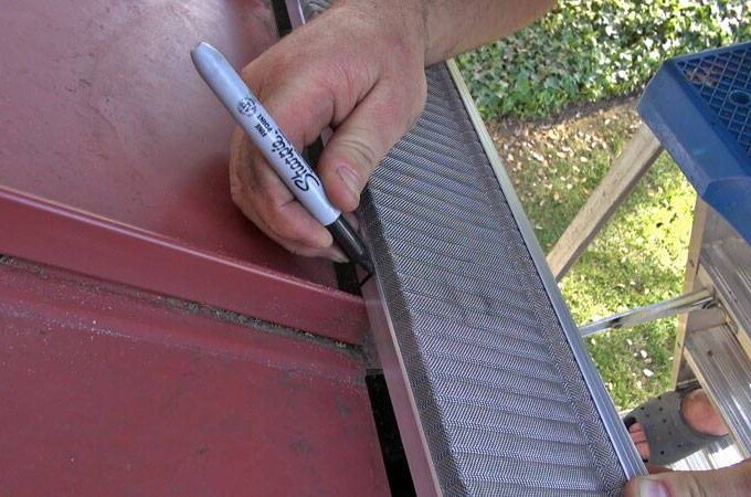 A person uses a black marker to mark a metal gutter guard while standing on a ladder next to a red roof, with grass and leaves visible below.