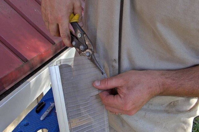 A person uses tin snips to cut a piece of metal mesh gutter guard while installing it along the edge of a roof.