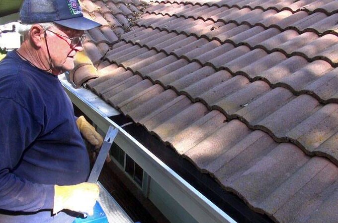 An older man wearing a cap, gloves, and glasses stands on a ladder cleaning leaves and debris from a house’s gutter beside a tiled roof, preparing for easy installation of Gutterglove.