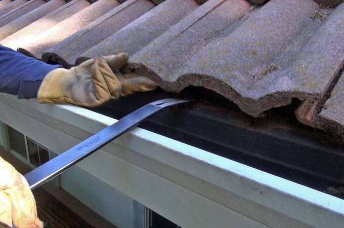 A person wearing work gloves uses a flat pry bar to lift a roof tile near the edge of a house, exposing the area above the rain gutter to install Easy On Gutter Guard.