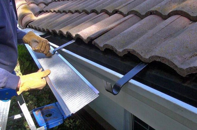 A person wearing gloves installs a Costco gutter guard with easy installation, placing a metal mesh guard over a house gutter beneath a tiled roof while standing on a ladder.