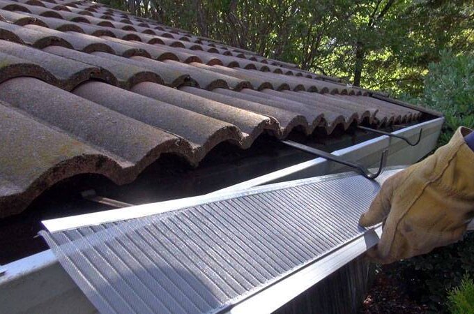 A person wearing work gloves easily installs a Gutterglove metal mesh gutter guard along the edge of a tiled roof, with trees visible in the background.