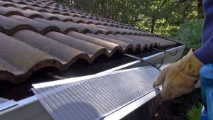 A person wearing work gloves installs a mesh gutter guard along the edge of a tiled roof to prevent debris from clogging the rain gutter. Trees and greenery are visible in the background.