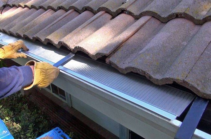 Person wearing gloves installs an Easy On Gutter Guard with stainless steel micro-mesh under roof tiles, preventing debris from entering the gutter below for lasting protection.