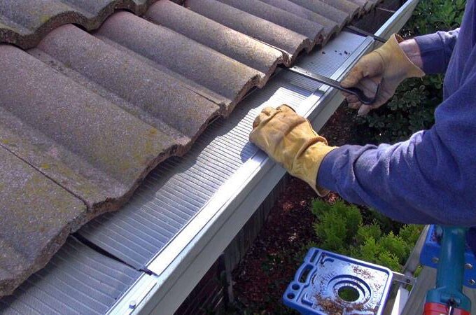 A person wearing gloves is installing a Gutterglove stainless steel micro-mesh gutter guard along the edge of a tiled roof, using a tool to secure it in place. Green shrubs and a toolbox are visible below the roof.